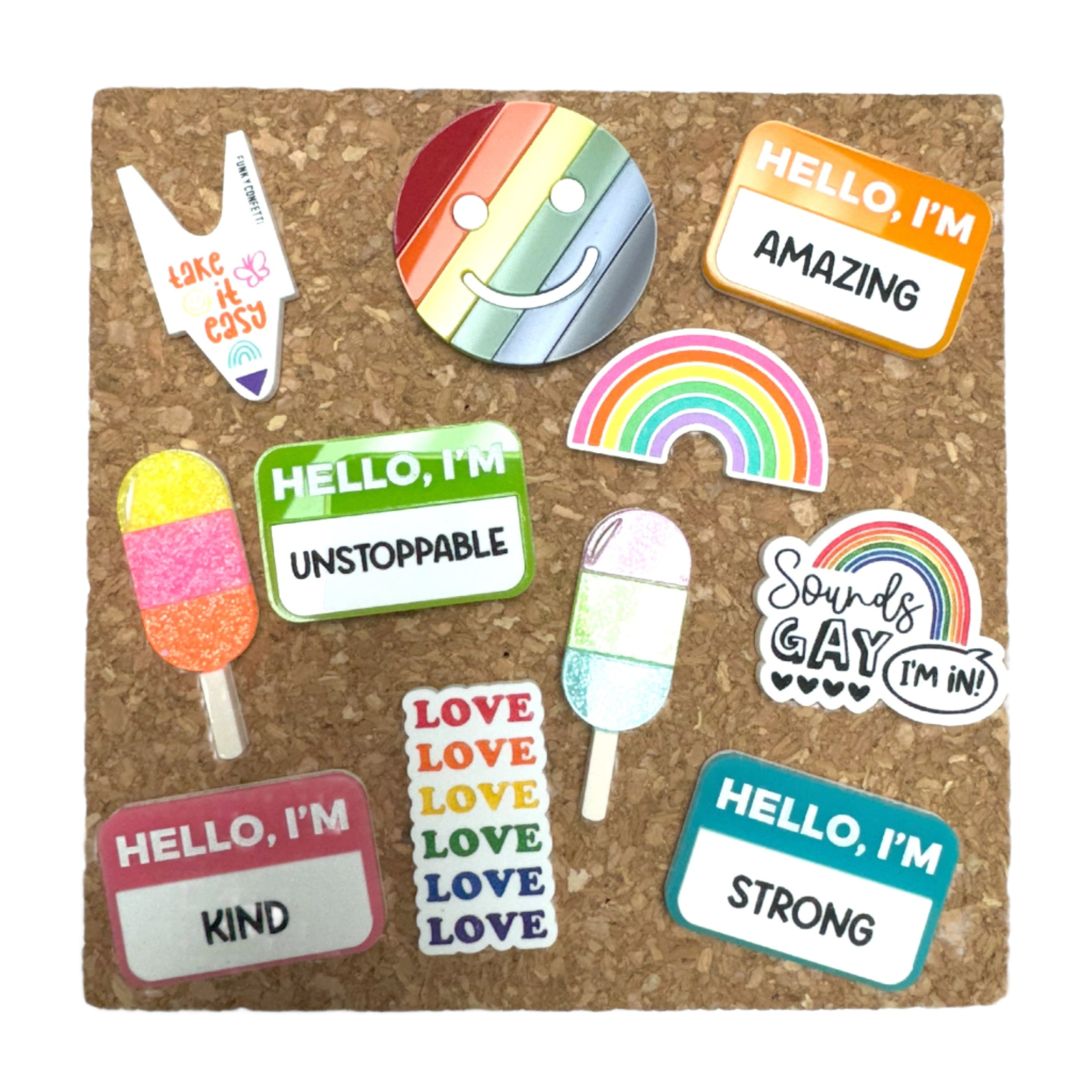 Corkboard display with assorted colorful pins: rainbow smiley face, rainbow arch, “Sounds Gay, I’m In!” cloud, stacked “LOVE” text, glitter popsicle pins, “take it easy” white V pin, and name-tag pins reading “HELLO, I’M AMAZING/UNSTOPPABLE/STRONG/KIND.