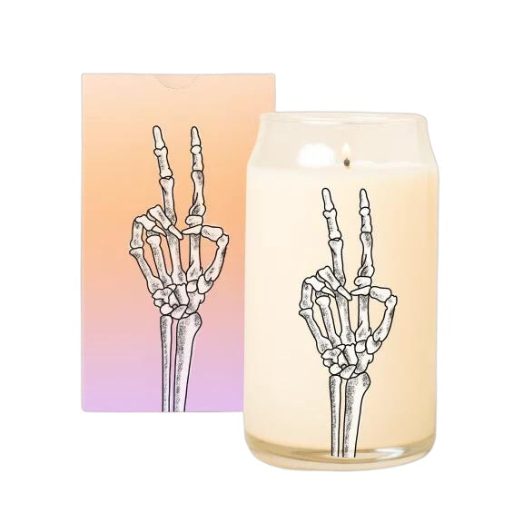 Can-shaped glass candle with black skeleton hand peace sign, shown beside gradient orange-to-purple gift box.