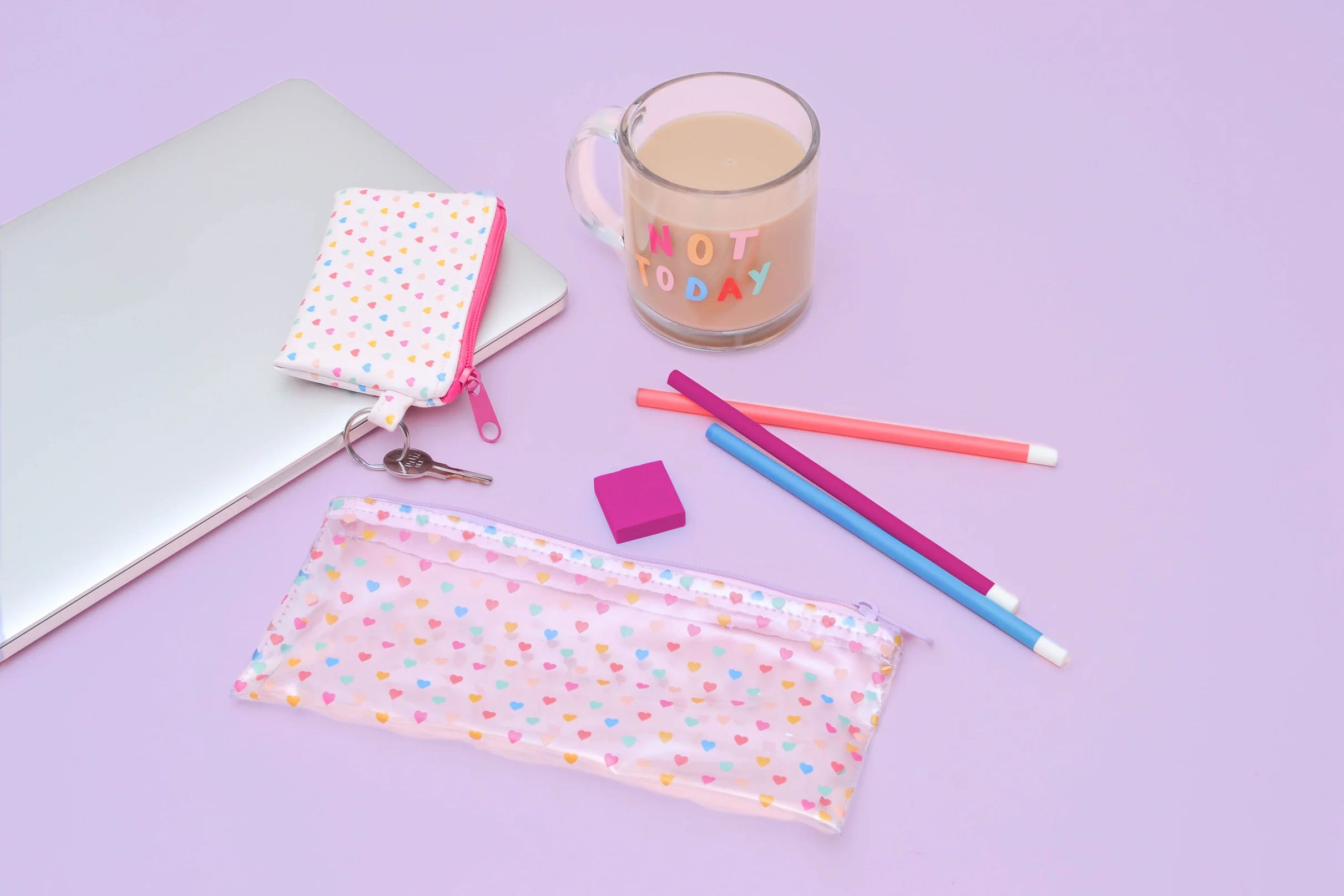 Lifestyle flat lay: transparent pencil pouch with tiny multicolor hearts and pink zipper, shown with a laptop, matching heart key pouch, pastel pencils, pink eraser, and a glass mug that reads “NOT TODAY” on a lavender background.