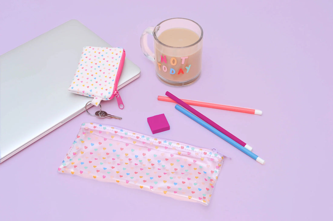 Lifestyle flat lay: transparent pencil pouch with tiny multicolor hearts and pink zipper, shown with a laptop, matching heart key pouch, pastel pencils, pink eraser, and a glass mug that reads “NOT TODAY” on a lavender background.