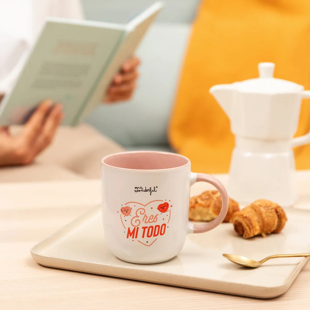Lifestyle: cream ceramic mug with blush interior and the phrase “Eres mi todo” in a heart, on a breakfast tray with croissants and a moka pot; person reading in the background.